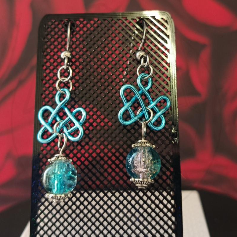Brincos pendentes com padrão entrelaçado azul e contas azuis transparentes em suporte preto perfurado.
