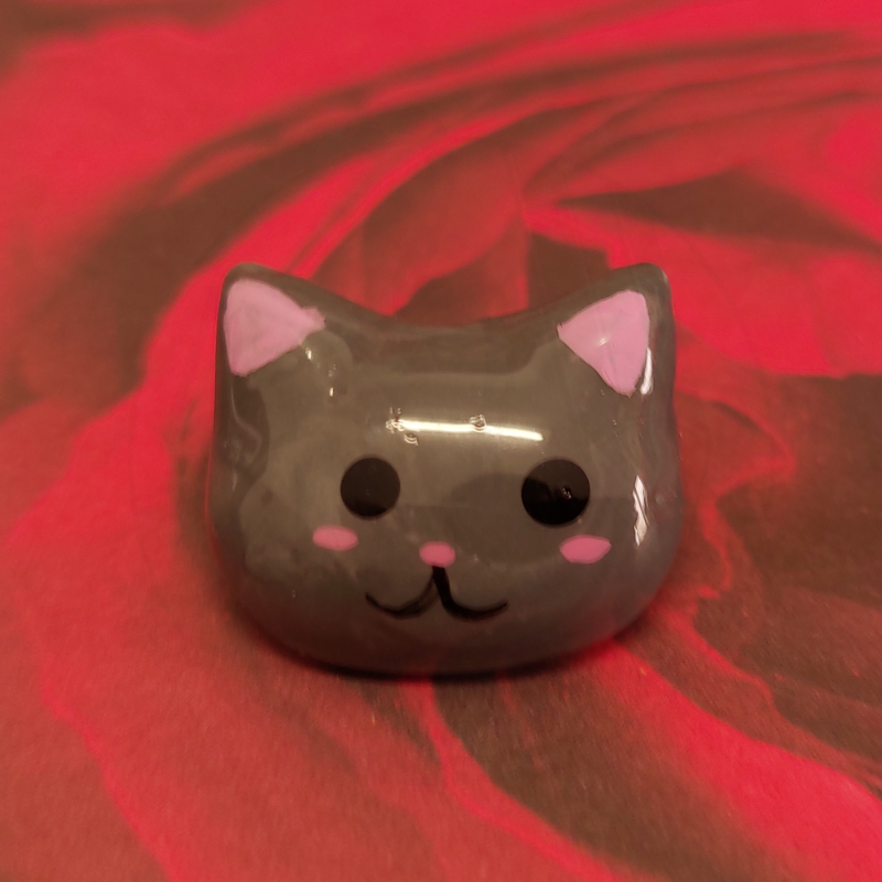 Figura decorativa cinzenta em forma de cabeça de gato com detalhes cor-de-rosa sobre fundo vermelho.