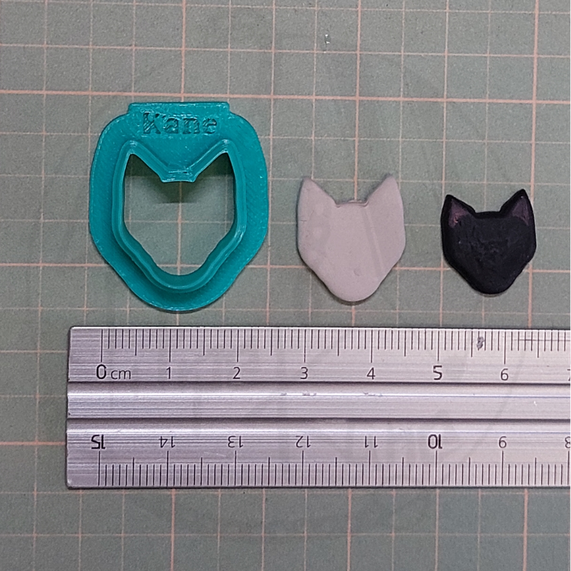 Cortador de biscoitos formato cabeça de gato azul, duas formas de massa cinzenta e preta, régua metálica sobre superfície quadriculada.