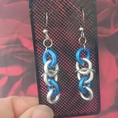 Brincos pendentes azuis e prateados em cartão preto com padrão de grelha.