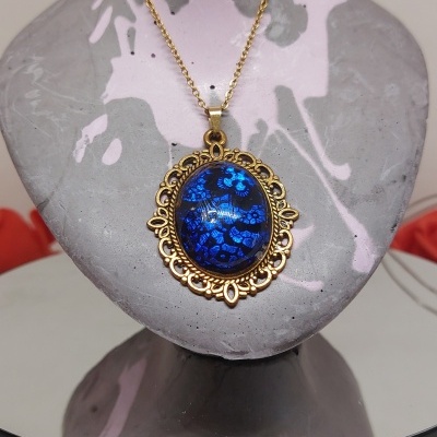 Colar dourado com pendente azul floral em busto cinza com manchas rosas sobre base preta com botões e cabo USB.