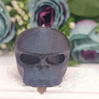 Objeto decorativo em forma de caveira escura com brilho azul sobre pano branco e flores ao fundo