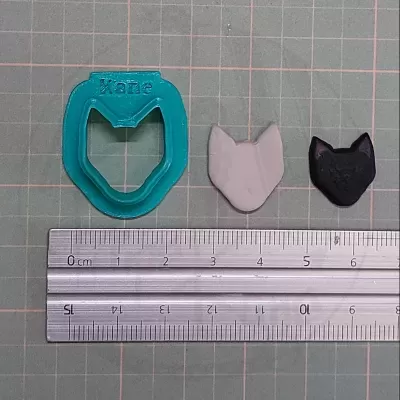 Cortador de biscoitos formato cabeça de gato azul, duas formas de massa cinzenta e preta, régua metálica sobre superfície quadriculada.