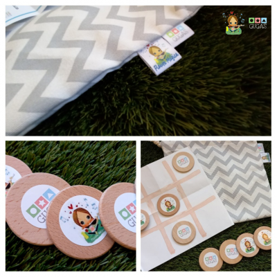 Jogo de tabuleiro com peças de madeira, saco de tecido cinzento com padrão chevron, etiquetas coloridas e folha de jogo sobre relva sintética.