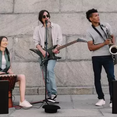 Três músicos de rua com djembê, guitarra elétrica e saxofone junto a caixas de som pretas em parede de pedra cinzenta.