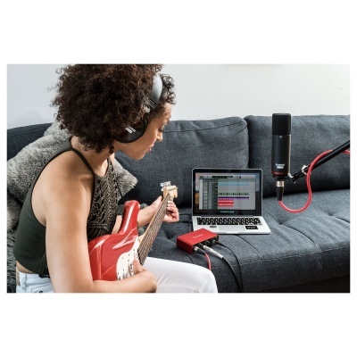 Mulher toca guitarra elétrica vermelha num sofá com laptop, interface áudio e microfone.