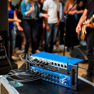 Equipamento áudio azul com cabos num ambiente de concerto ao fundo.