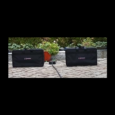 Dois altifalantes pretos DYNAMO no chão de calçada com plantas atrás
