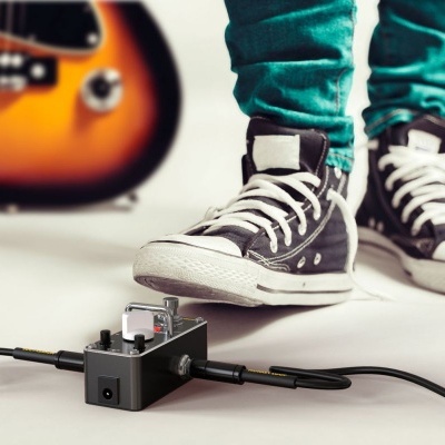Pedal de efeito de guitarra preto com botões prateados ligado a cabos, com pessoa de ténis e calças verdes e guitarra acústica no fundo.