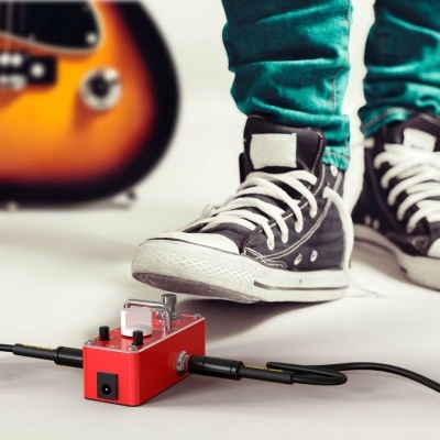 Pedal de efeito de guitarra vermelho com botões metálicos ligado a cabos, sapatilhas pretas estilo Converse e guitarra elétrica ao fundo
