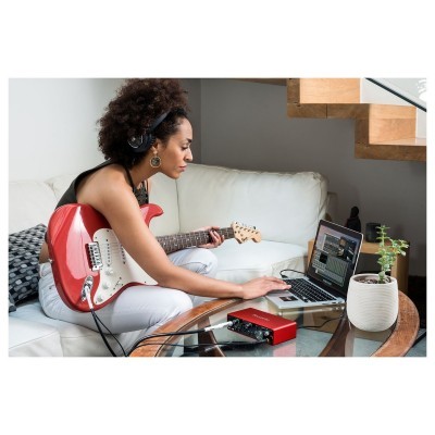 Mulher sentada no sofá a tocar guitarra elétrica vermelha com computador e interface de áudio