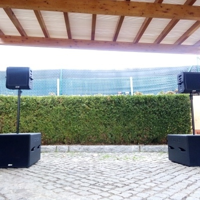 Dois altifalantes pretos em suportes elevados, chão de pedra e cerca verde ao fundo