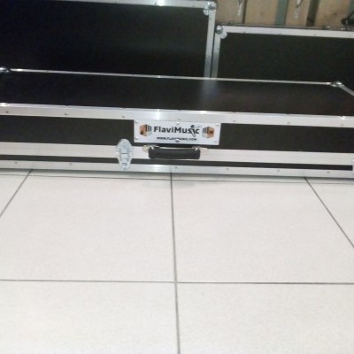 Mala rígida preta com acabamentos metálicos e etiqueta FLAVIMUSIC colocada em chão de azulejos claros