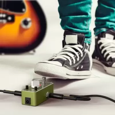 Pedal de efeitos verde metálico no chão com cabos ligado, guitarra elétrica e pés com ténis e calças verdes ao fundo