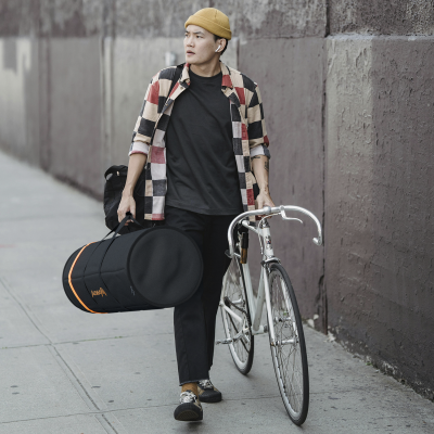 Homem com bicicleta e bolsa para instrumento musical numa rua urbana, vestido casual com camisa xadrez e boné bege.