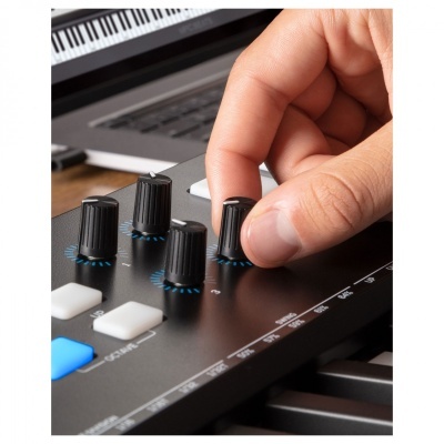 Mão ajustando botão num teclado controlador MIDI preto com teclado de piano e portátil ao fundo