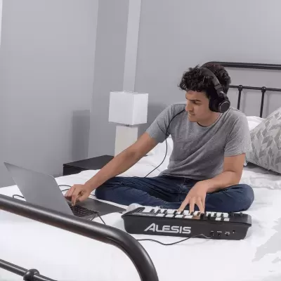 Homem sentado na cama a usar controlador MIDI ALESIS com portátil e auscultadores.