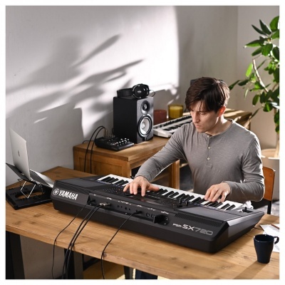 Teclado musical Yamaha SX7AD preto numa mesa com homem a tocar e equipamento de som e informática