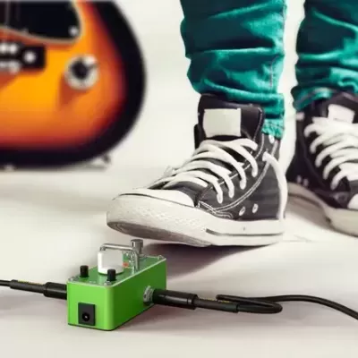 Pedal de efeitos verde no chão com pessoa e guitarra no fundo