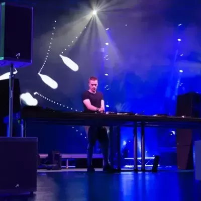 DJ em palco com mesa de mixagem e colunas grandes pretas sob luzes azuis