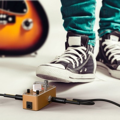 Pedal de efeito musical dourado em chão branco com pessoa e guitarra ao fundo.