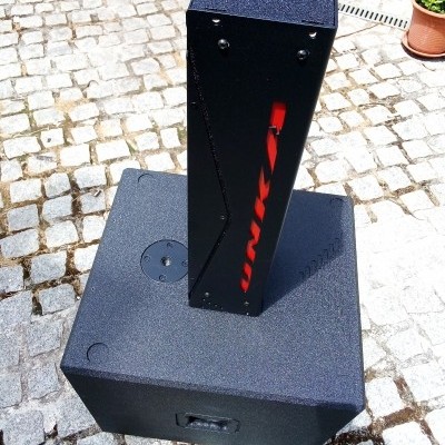Sistema de som portátil preto com palavra UNIKA em vermelho numa coluna vertical sobre piso de pedras irregulares