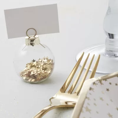 Ornamento esférico transparente com confetes dourados e suporte para cartão junto a talheres dourados e prato com estrelas.