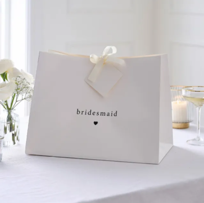 Saco de papel branco com texto bridesmaid e laço creme sobre mesa branca com flores, vela e copo