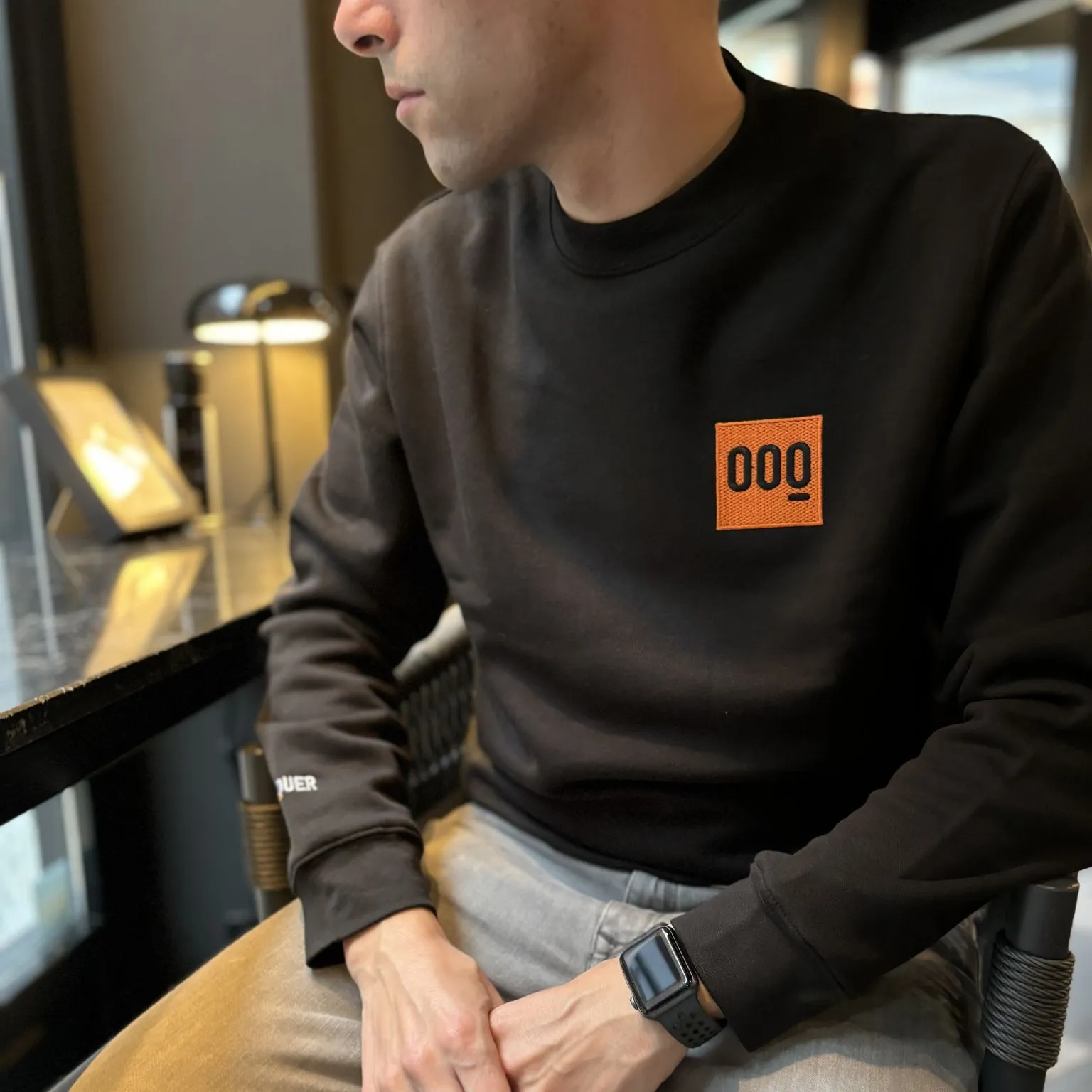 Sweatshirt preta com logo laranja e preto 000 usada por pessoa sentada junto a mesa