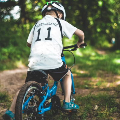 Criança de costas a andar de bicicleta azul num trilho de terra com t-shirt branca e capacete.