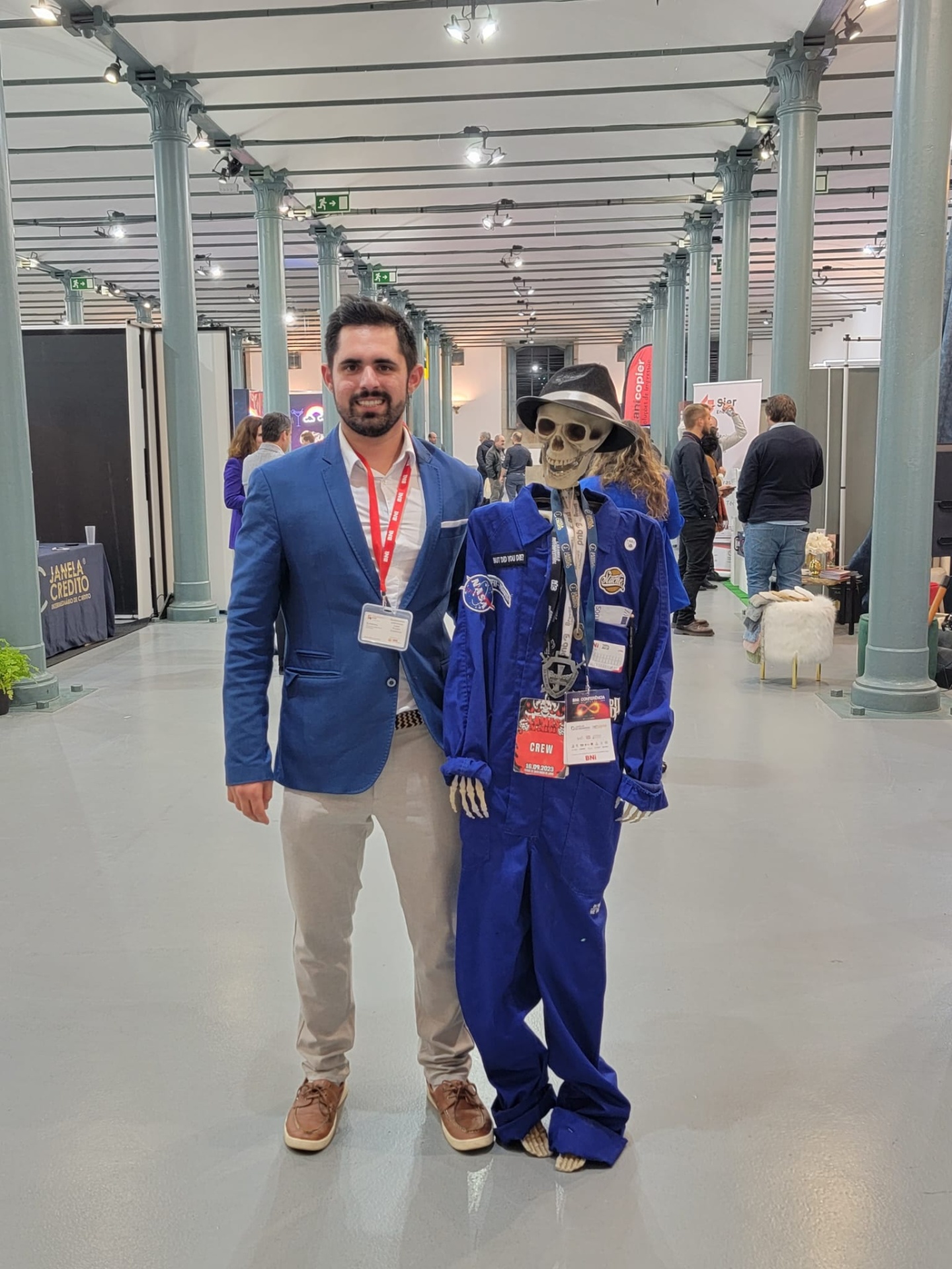 Homem ao lado de esqueleto vestido com macacão azul e chapéus em espaço interior