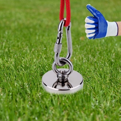 Imã metálico redondo com corda e mosquetão na relva, mão com luva a fazer sinal de positivo