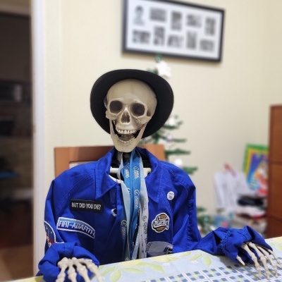 Esqueleto com camisa azul e chapéu preto sentado à mesa numa sala decorada