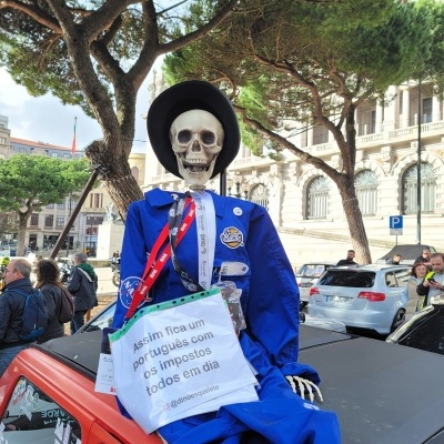 Boneco esqueleto vestido com fato azul e chapéu preto no tejadilho de carro vermelho com cartaz sobre impostos