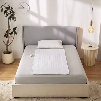 Cama estofada bege claro com colchão cinza e almofada branca em quarto minimalista com planta, mesa lateral e relógio