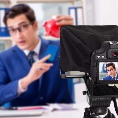 Câmara digital preta com ecrã traseiro focado num homem de fato azul a apontar para um cofrinho