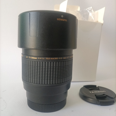 Objetiva fotográfica Tamron preta com tampa e caixa branca ao fundo