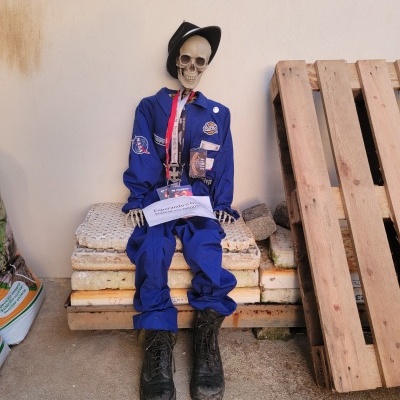 Figura esqueleto com macacão azul e botas pretas sentada em colchões ao lado de palete de madeira.