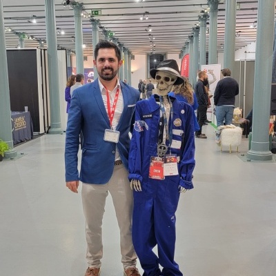 Homem ao lado de esqueleto vestido com macacão azul e chapéus em espaço interior