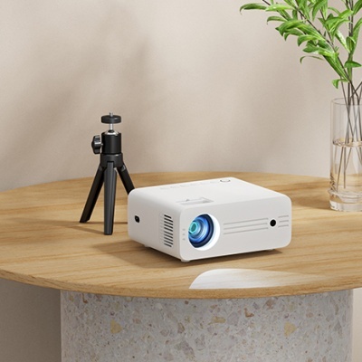 Projetor branco numa mesa de madeira ao lado de mini tripé e vaso com folhas