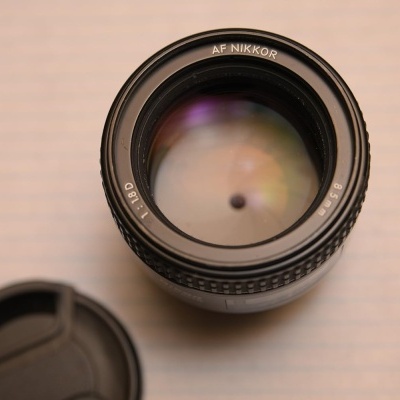 Lente fotográfica AF NIKKOR 50mm 1:1.8D preta sobre superfície branca com linhas e tampa ao lado