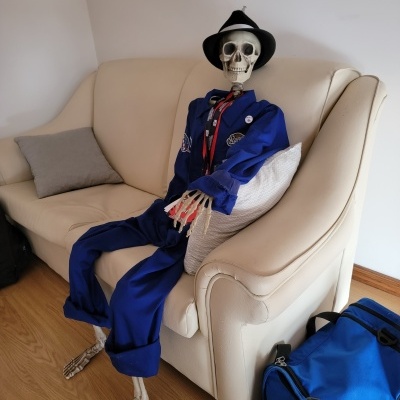 Esqueleto vestido com fato azul sentado num sofá branco com mochila azul ao lado