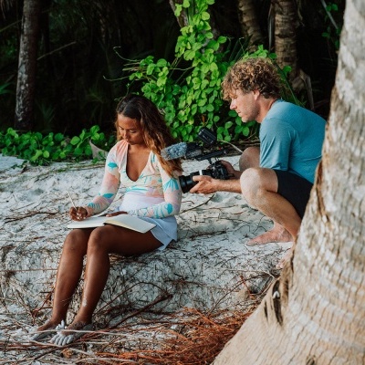 Mulher sentada na praia a escrever com homem a filmar com câmara de vídeo