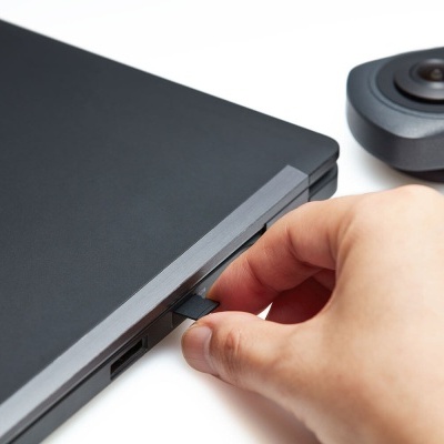 Mão a inserir pen USB preta num portátil preto com uma câmara preta ao fundo