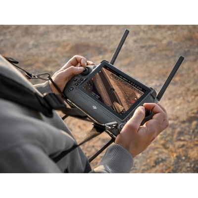 Comando remoto DJI com ecrã e duas antenas, mãos segurando, terreno arenoso ao fundo