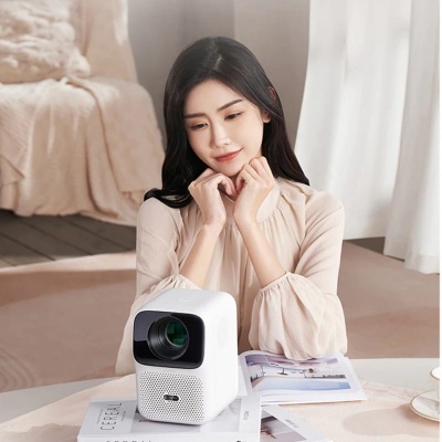 Projetor digital branco com lente preta numa mesa branca com revistas, mulher sentada ao fundo