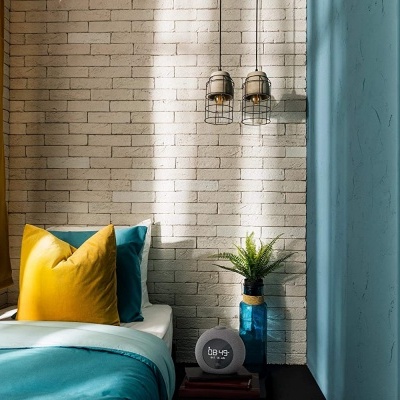 Quarto com parede branca de tijolo, cortinas amarelas, cama com almofadas amarelas e azuis e mesa de cabeceira com planta e relógio preto