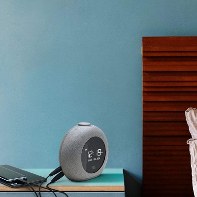 Relógio digital cinza com ecrã preto ao lado de telefone e cama