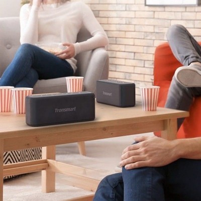 Coluna de som portátil preta Tronsmart numa mesa de madeira com copos de papel e pessoas sentadas em sofás ao fundo