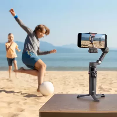 Gimbal DJI cinzento com smartphone a filmar crianças a jogar futebol na praia